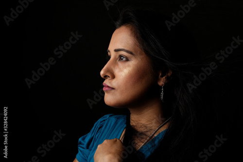 Close up face portrait of a beautiful Indian Bengali brunette woman in light and shadow before a black copy space background wearing a blue top. Indian lifestyle and fashion portrait