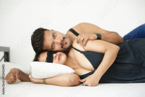 Attractive Indian Bengali brunette couple sharing intimate moments wearing black dress lying on bed in a white bedroom