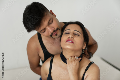 Attractive Indian Bengali brunette couple seductive foreplay in black dress in white bedroom while the man trying to strangle the throat of the girl from her back.  Indian lifestyle domestic violence photo