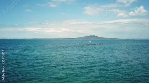 aerial drone shot, flying over a person fishing on the reef into a fishing boat in the middle of the sea with Rangitoto island as background in New Zealand. photo