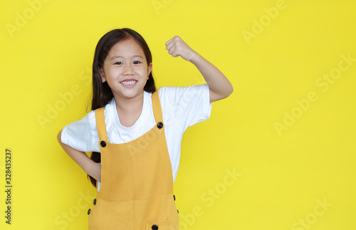 Happy asian little child girl showing his muscle with looking camera isolated on yellow background with copy space. Strong kid in dungarees with good health concept photo