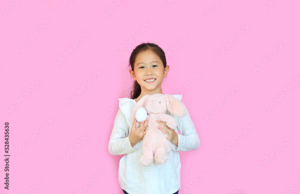 Pretty asian little child girl holding pink fluffy rabbit doll isolated on pink background with looking camera. Happy Kid on Easter. Pretty asian little child girl holding pink fluffy rabbit doll isolated on pink background with looking camera. Happy Kid on Easter.
