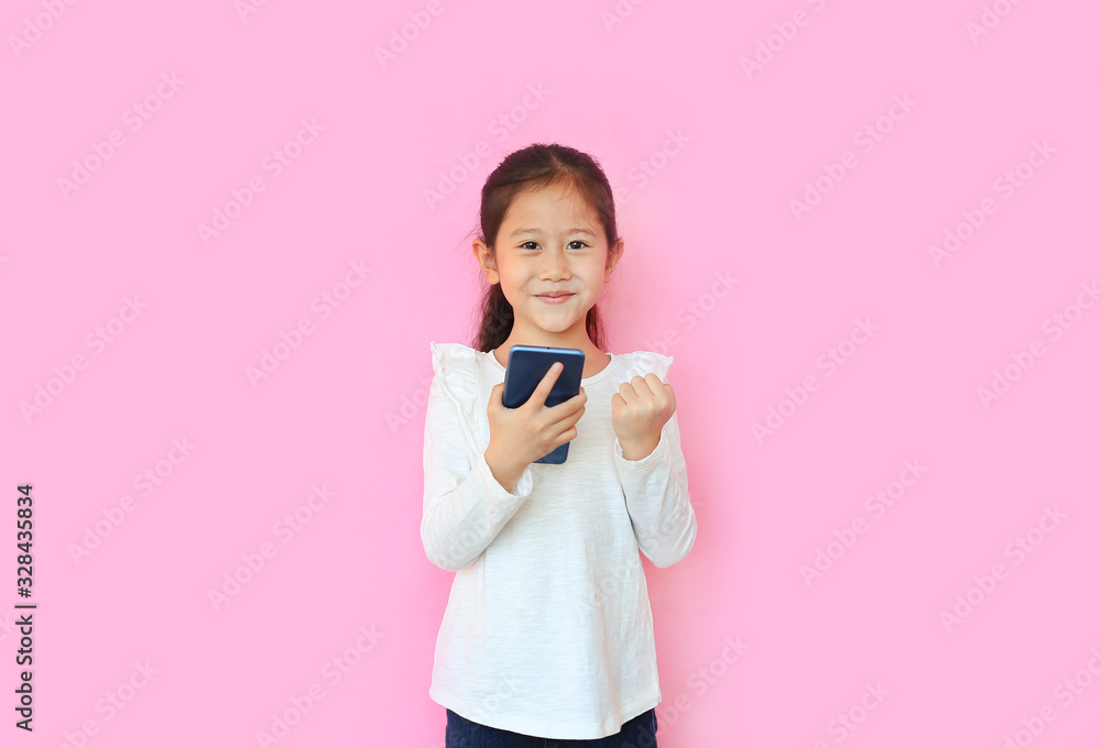 Happy asian little child girl watching her smartphone and expression hand fight sign over pink background. Kid smiling and enjoy while playing cellphone. Happy asian little child girl watching her smartphone and expression hand fight sign over pink background. Kid smiling and enjoy while playing cellphone.