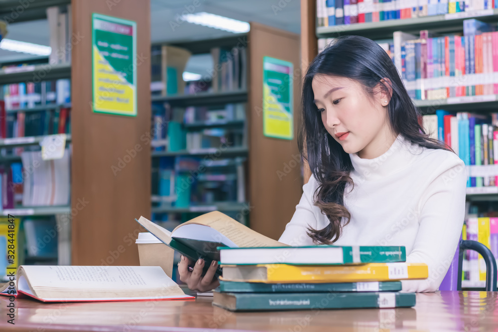 Young beautiful Asian female student portrait sitting and concentrate studying or reading textbook in university library for exams or test with book shelves background. Education and learning concept Young beautiful Asian female student portrait sitting and concentrate studying or reading textbook in university library for exams or test with book shelves background. Education and learning concept