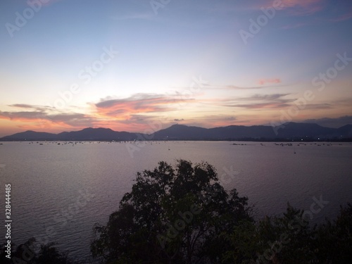 Seascape and sunset. Glow and ray of sun down at the cloudy sky.  Hills of island on the background on the horizon  silhouette of tree crowns on the foreground. Panorama of water in twilight evening