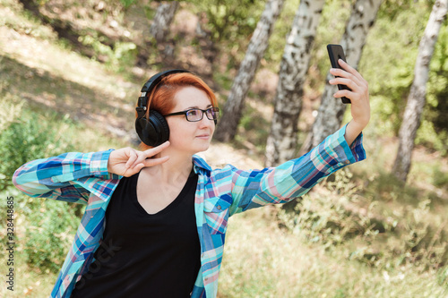 Young ginger woman makes selfie in park. Cute redhead girl in glasses and headphones with smartphone in forest.
