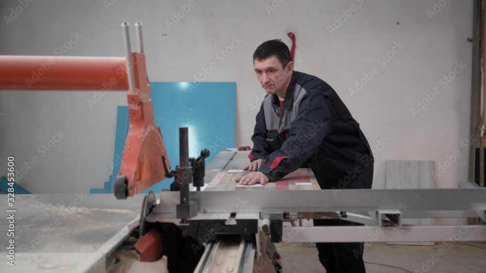 An adult male worker in working clothes works in a carpentry workshop. He saws the board on an electric saw. An adult male worker in working clothes works in a carpentry workshop. He saws the board on an electric saw.