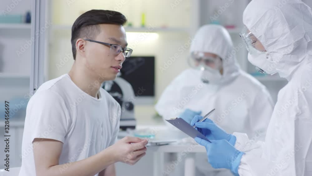Tracking shot of sick male Asian patient in glasses describing his symptoms to unrecognizable doctor in disposable coveralls, safety goggles and gloves Tracking shot of sick male Asian patient in glasses describing his symptoms to unrecognizable doctor in disposable coveralls, safety goggles and gloves