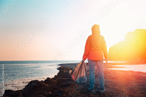 A young Caucasian female volunteer in a jacket poses with a large garbage bag and a plastic bottle in her hands. Rear view. In the background  the sky and the sea. Copy space