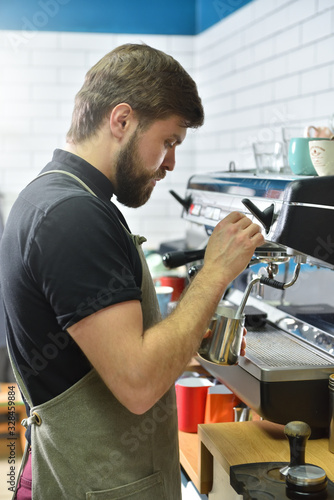 A bartender with a beard is making coffee on a coffee machine and whipping milk with steam. Small business concept