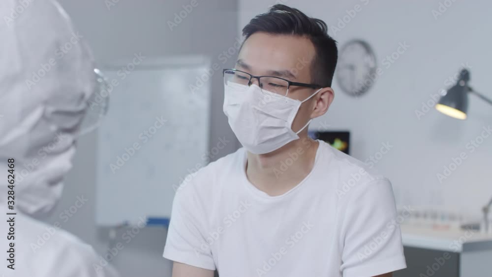 PAN shot of unrecognizable doctor in protective suit and goggles listening to potentially contagious male Asian patient in face mask describing his symptoms PAN shot of unrecognizable doctor in protective suit and goggles listening to potentially contagious male Asian patient in face mask describing his symptoms