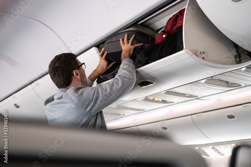Tableau sur toile Young man putting luggage into overhead locker on airplane