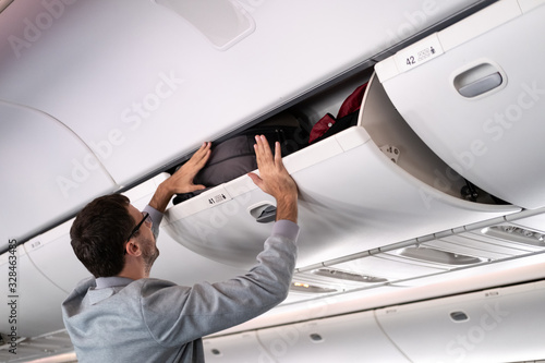 Young man putting luggage into overhead locker on airplane. Traveler placing carry on bag in overhead compartment photo