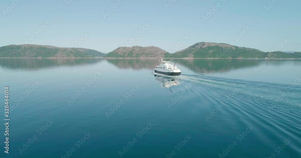 Wide drone shot with parallax tilt motion revealing small passenger ferry sailing on the Norwegian coast in calm and clear turquoise water reflecting the ship, near the arctic circle in Norway. Wide drone shot with parallax tilt motion revealing small passenger ferry sailing on the Norwegian coast in calm and clear turquoise water reflecting the ship, near the arctic circle in Norway.