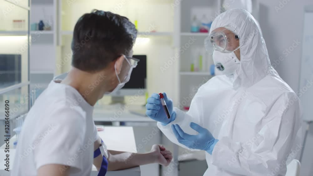 Medium shot of unrecognizable doctor in disposable coveralls, goggles, respiratory mask and gloves holding tube with blood sample and explaining something to sick patient Medium shot of unrecognizable doctor in disposable coveralls, goggles, respiratory mask and gloves holding tube with blood sample and explaining something to sick patient