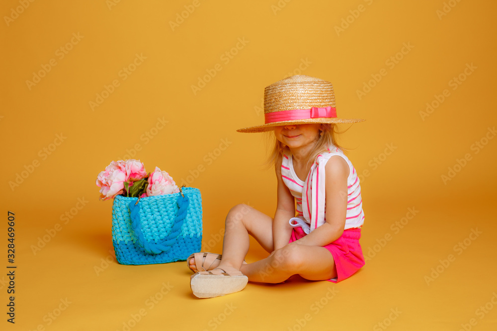 a little girl in summer clothes and a bag of flowers hid under a hat on a yellow background. The concept of summer vacation a little girl in summer clothes and a bag of flowers hid under a hat on a yellow background. The concept of summer vacation