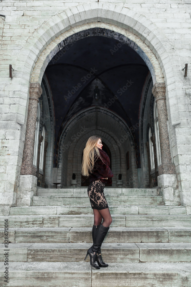 Young girl dressed fur coat and dress standing and look straight on the stairs of church. Old church on the background.