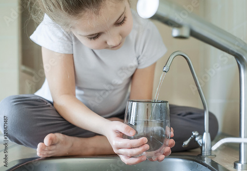Little girl open a water tap with her hand holding a transparent glass. Kitchen faucet. Filling cup beverage. Pouring fresh drink. Hydration. Healthcare. Healthy lifestyle. World water day