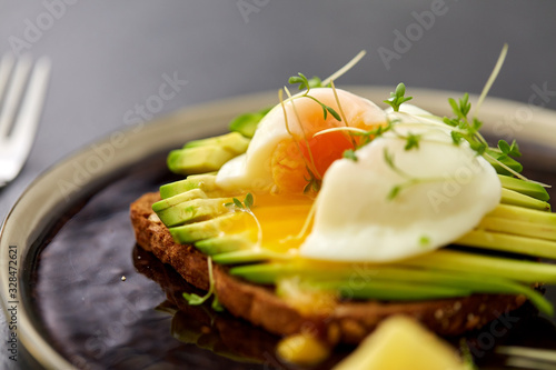 food, eating and breakfast concept - toast bread with sliced avocado, pouched egg and greens on ceramic plate