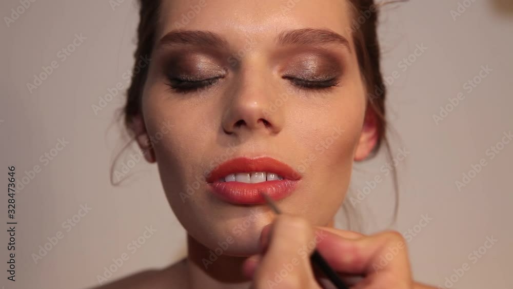gorgeous girl looking at camera and a make up artist applying lipstick with a brush on her lips. gorgeous girl looking at camera and a make up artist applying lipstick with a brush on her lips.