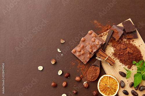 sweets, confectionery and culinary concept - chocolate with hazelnuts, cocoa beans, and powder with dry orange and cinnamon on wooden board with spoon on brown background