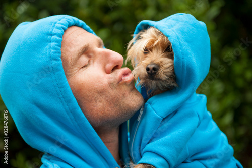 Portrait of dog owner kissing a furry friend in matching blue hoodies outdoors in bright green park background