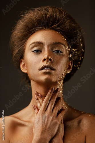Fototapeta Fashion art portrait of model girl with holiday golden shiny professional makeup. beaty woman with golden sparkles on skin and hair on dark background. Gold glowing skin.