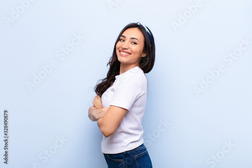 young latin pretty woman smiling gleefully, feeling happy, satisfied and relaxed, with crossed arms and looking to the side against flat wall