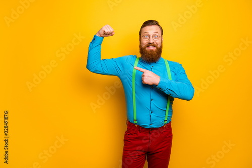 Look. Photo of attractive funny toothy guy raise fist arm indicating finger presenting perfect shape biceps wear blue shirt green suspenders red pants isolated yellow color background