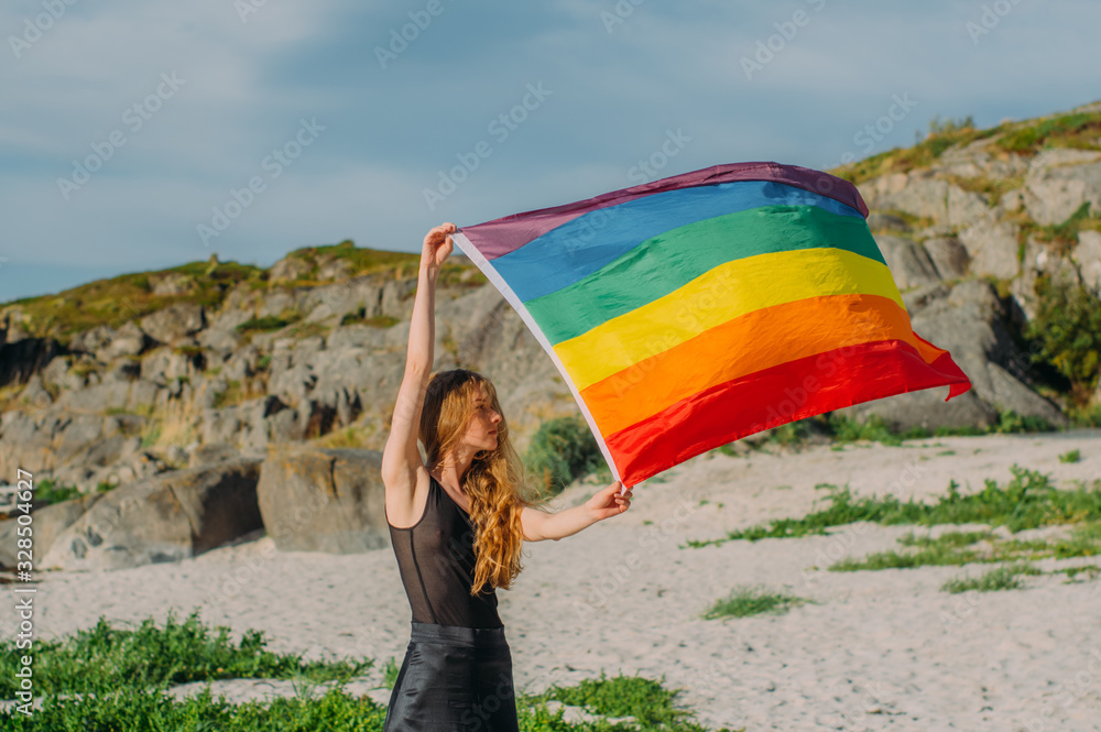 Young blond girl holding  LGBTQI flag  walking in the day on the white beach near the sea