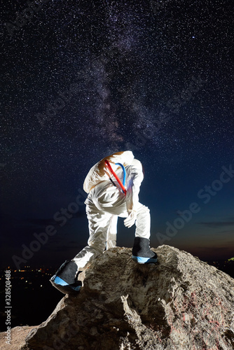 Back view of space traveler reaching top of rocky mountain under mesmerizing night sky with stars  Milky way. Male cosmonaut in space suit exploring new planet. Concept of space travel  cosmonautics