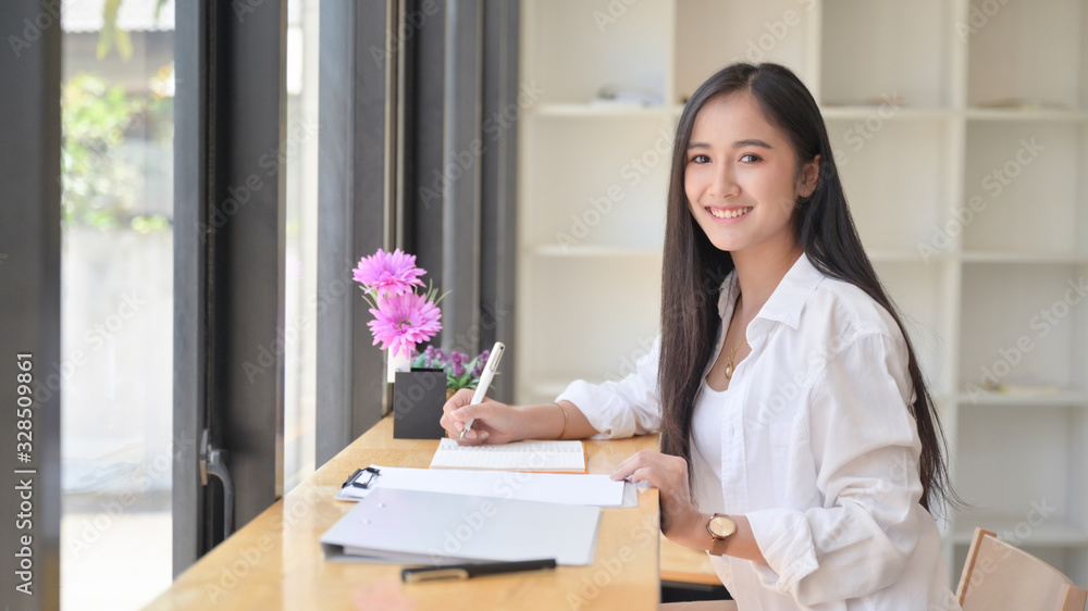 Asian woman wearing a white dress holding a pen to write notes in a comfortable office. She looked at the camera and smiled.