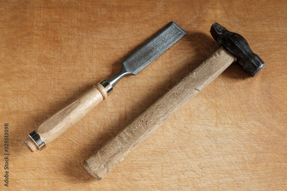 One chisel with a wooden handle and an old hammer with a wooden handle lie diagonally on a wooden table. One chisel with a wooden handle and an old hammer with a wooden handle lie diagonally on a wooden table.