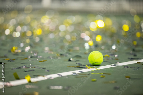 A blank yellow tennis ball next to the side line on a green tennis court with sparkling and shiny confetti after a winning match.