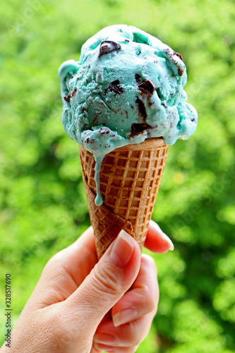 Woman's Hand Holding a Cone of Melting Mint Chocolate Chip Ice Cream on Blurry Green Garden Background