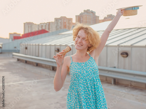 young happy cheerful curly redhead woman in straw hat, blue sundress eating ice cream on skyscraper roof (skyroof). Fun, urban, modern, roof, city, summer, fashion, youth concept photo