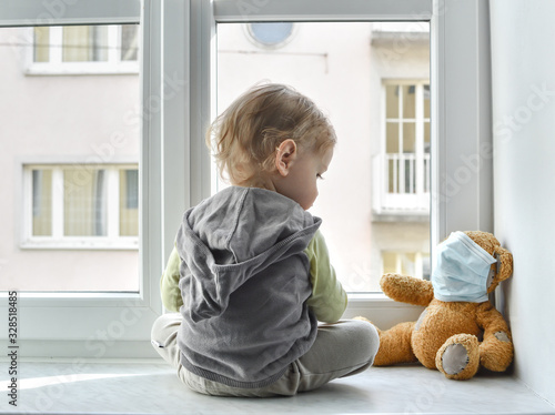 Child in home quarantine standing at the window with his sick teddy bear wearing a medical mask against viruses during coronavirus and flu outbreak. Children and illness COVID-2019 disease concept