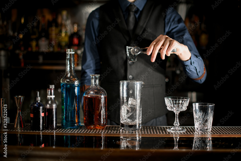 Bartender is preparing alcoholic cocktail with ice in a glass behind the bar.