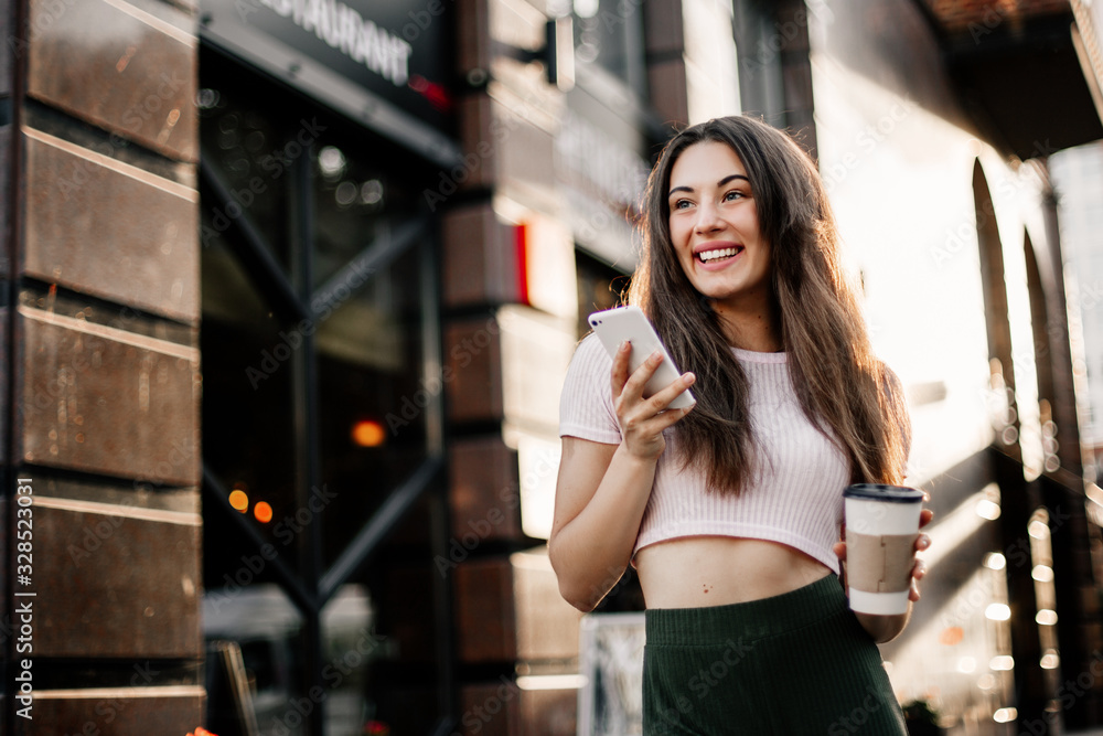 Smiling woman using phone and drink coffee on the street in summer day.