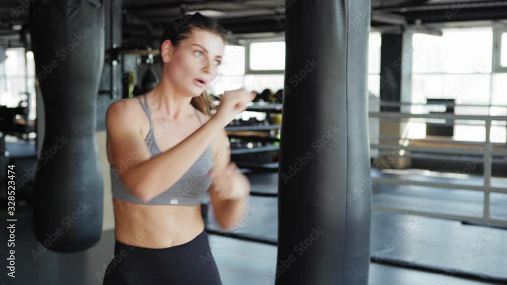 Tilt up side view waist up shot of fit young woman in crop top practicing boxing punches standing among heavy bags in gym Tilt up side view waist up shot of fit young woman in crop top practicing boxing punches standing among heavy bags in gym
