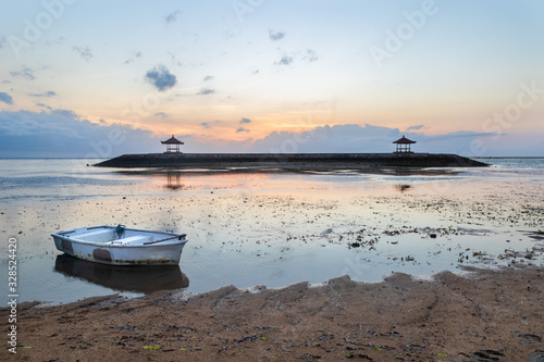 Sunrise at Sanur beach, in Bali, Indonesia. White boat near shore in the low tide. Twin pagodas sit on raised ground beyond. Clouds, colorful sky in the distance. 