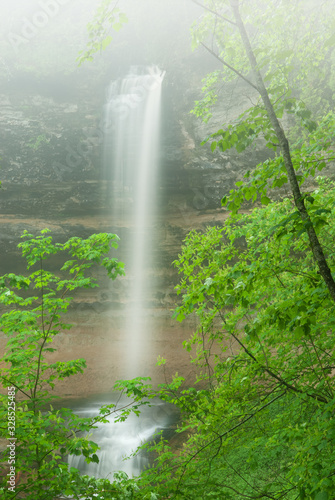 Foggy summer landscape of Munising Falls captured with motion blur and framed by branches and leaves, Michigan's Upper Peninsula, USA