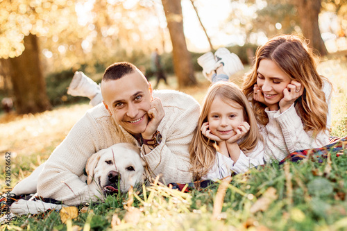 Happy family is having fun with dog labrador are lying on green grass in park. © zadorozhna