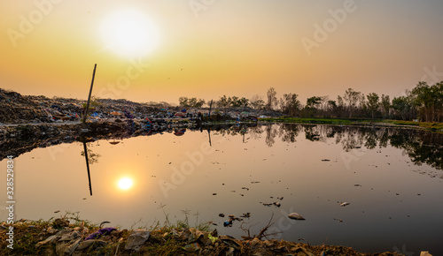 Polluted water and mountain large garbage pile and pollution at the sun is setting in the background