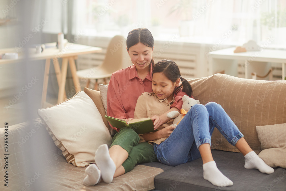 Horizontal shot of beautiful Asian woman relaxing on sofa with her lovely daughter reading interesting novel Horizontal shot of beautiful Asian woman relaxing on sofa with her lovely daughter reading interesting novel