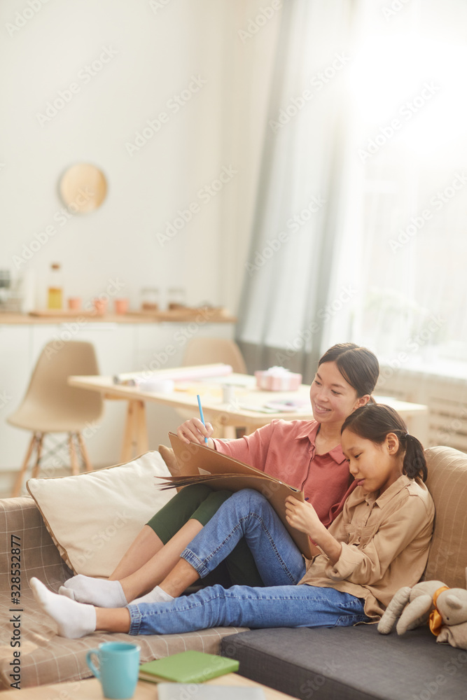 Asian woman and her daughter spending time together relaxing on sofa and drawing something on paper, vertical shot