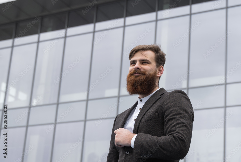 Portrait of an handsome businessman wearing jacket over office building in financial district outdoors. Young bearded Businessman outdoor. Successful entrepreneur dressed in formal wear.