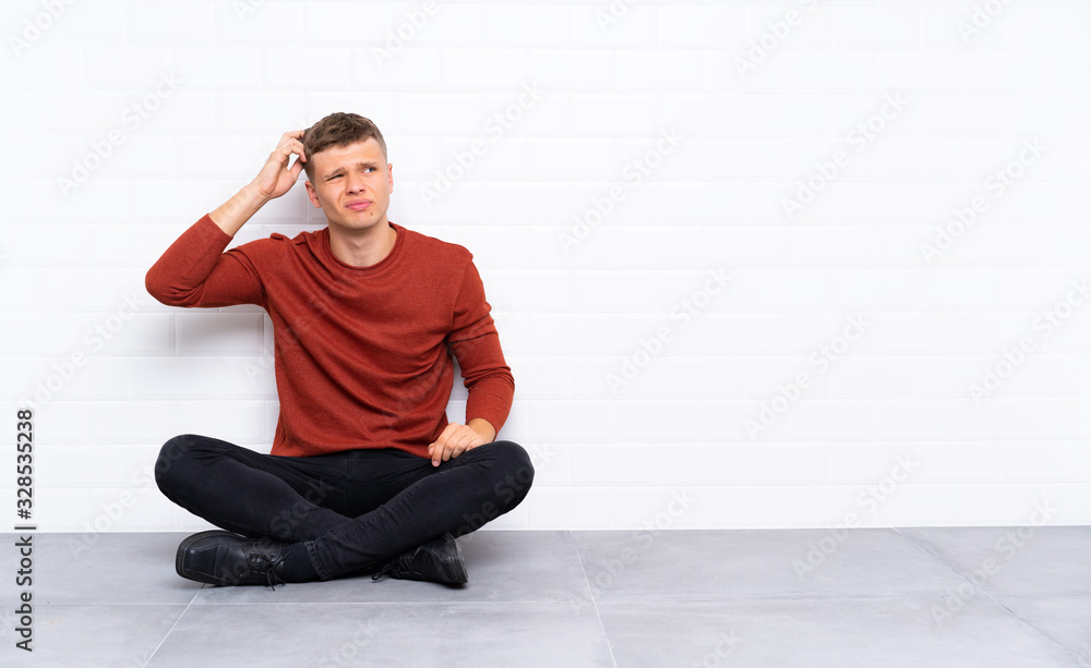 Young handsome man sitting on the floor having doubts and with confuse face expression Young handsome man sitting on the floor having doubts and with confuse face expression
