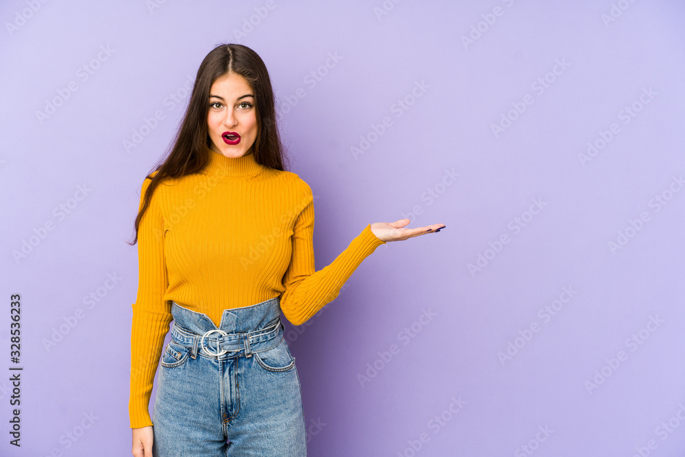 Young caucasian woman isolated on purple background impressed holding copy space on palm. Young caucasian woman isolated on purple background impressed holding copy space on palm.