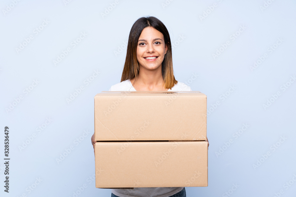 Young woman over isolated blue background holding a box to move it to another site Young woman over isolated blue background holding a box to move it to another site